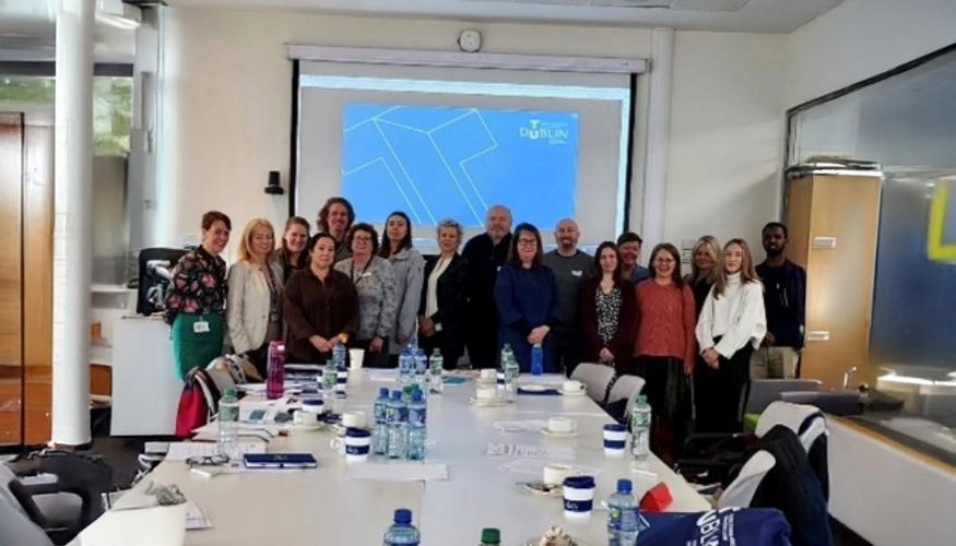 Group of adults posing, standing in front of table and chairs with TU Dublin screen in background