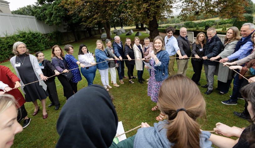 Lots of people in a circle outside on grass holding rope with a speaker in the centre.