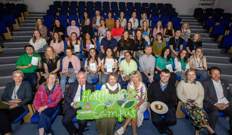 group of people sitting in lecture theatre facing camera for group photo. 3 hold healthy campus banner and one holds plate of food