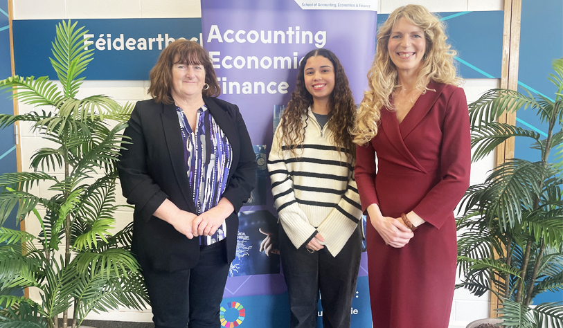 Three women standing posed for a photo in front of plants and a lilac TU Dublin school banner saying accounting Economics and Finance