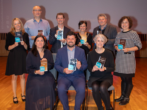 An image of 12 people, 6 standing and 6 standing in front of a screen holding their awards