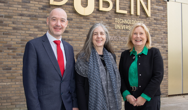 A group of people smiling in front of a brass TU Dublin logo on the University's campus in Grangegorman