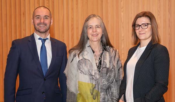 A photo of three people standing in front of wooden panels and smiling